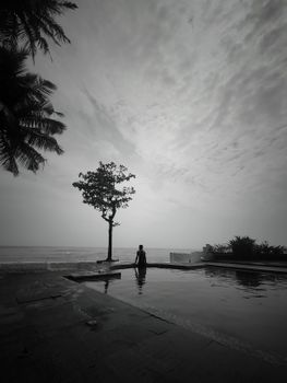 Silhouette of a person near a pool with a tree against a gray sky, creating a tranquil sunset scene in Kannur, India.