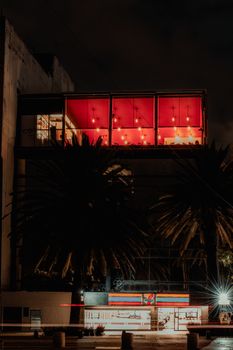Night view of a brightly lit building facade with palm trees, set against a dark sky.