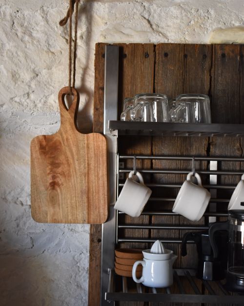 Free A rustic kitchen scene featuring a wooden cutting board, mugs, and glassware on a metal rack against a stone wall. Stock Photo