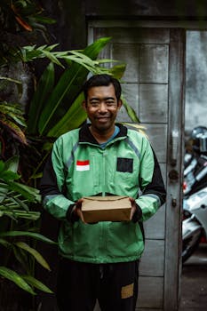 A cheerful delivery person in a green jacket holds a package, standing outdoors surrounded by plants.