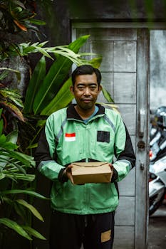 Smiling deliveryman in green jacket holds package, standing outdoors with lush greenery.