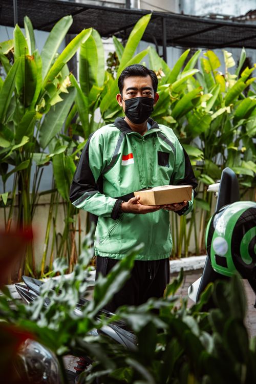 Free Delivery rider wearing mask holding food container in urban setting with greenery. Stock Photo