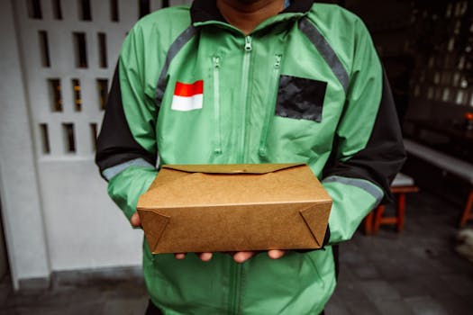 A delivery person in a green jacket holding a cardboard package outdoors.