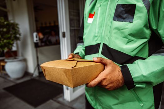 A deliveryman in a green jacket holding an eco-friendly food package outside a restaurant.