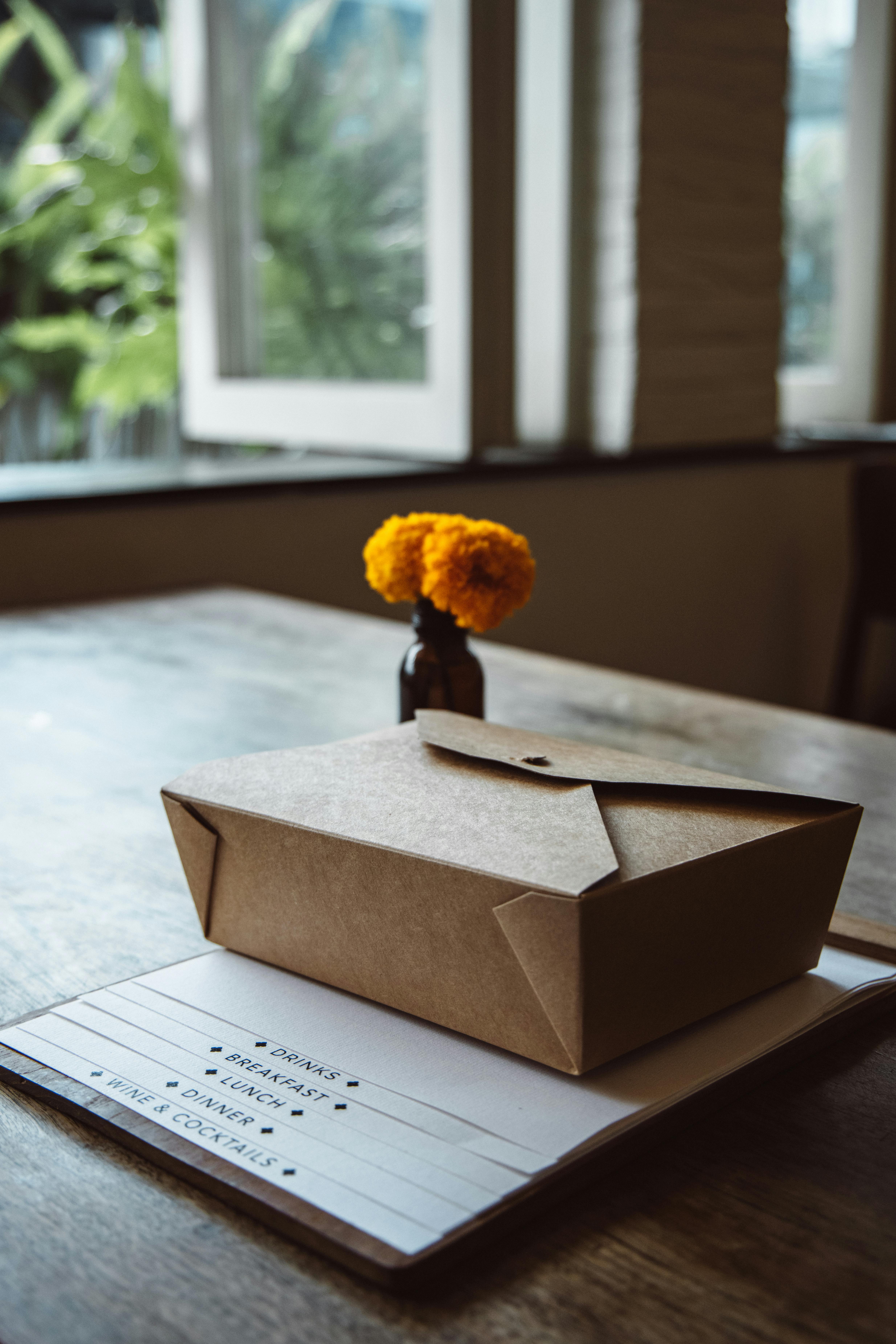 A brown cardboard lunchbox on a table with a flower in the background, creating a cozy indoor scene.
