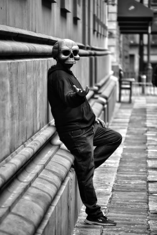 Grayscale Photo of a Person in a Skull Mask Leaning on a Wall