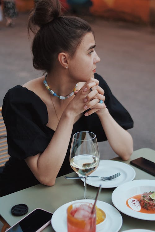 Free Woman Sitting Behind a Table in a Restaurant with Dishes and Wine in front of Her Stock Photo