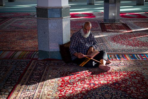 Free An elderly man with a beard reads in a mosque, surrounded by vibrant Persian carpets. Stock Photo