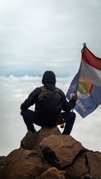 A man holding a flag sits on a rocky mountain peak, overlooking the clouds in Indonesia. - Fotografer Sidoarjo