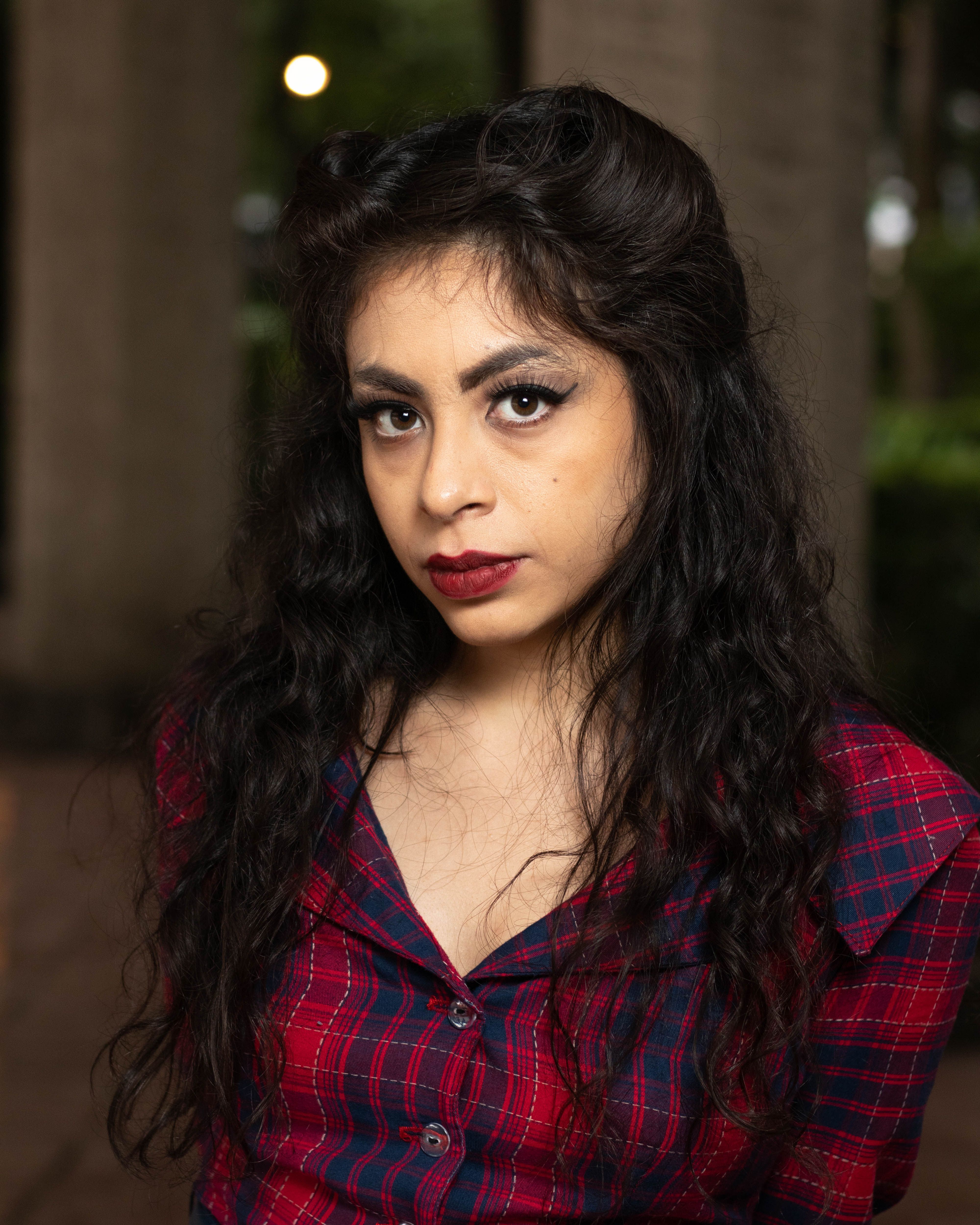 Close-up portrait of a woman with curly hair and red lips, wearing a plaid shirt, outdoors.