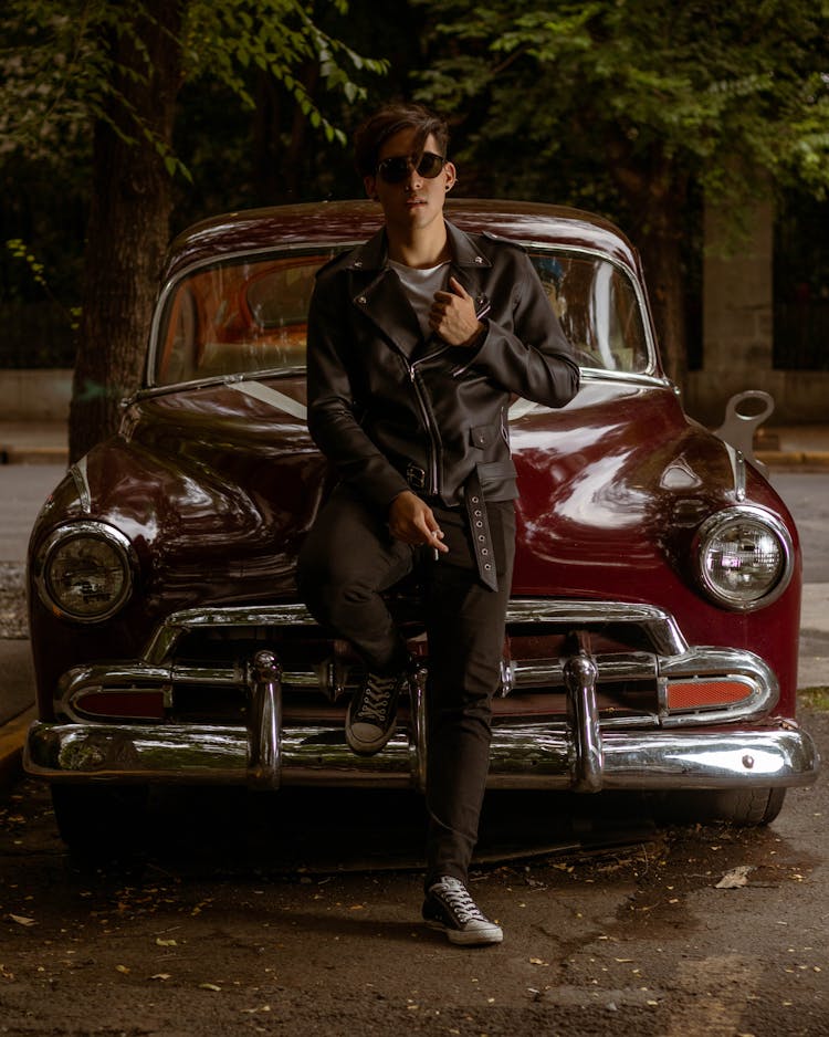 Man In Black Leather Jacket And Black Pants Leaning On A Vintage Car