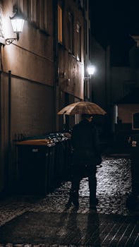 A lone man walks through a dark, rainy alleyway at night, holding an umbrella.