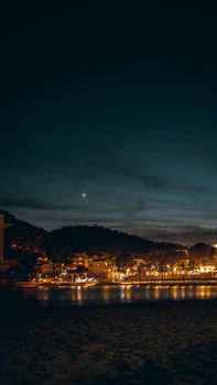A tranquil seaside town illuminated against the night sky, reflecting lights on serene waters.