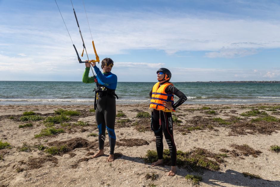 Two men in wetsuits preparing for a kitesurfing session on a sandy beach.