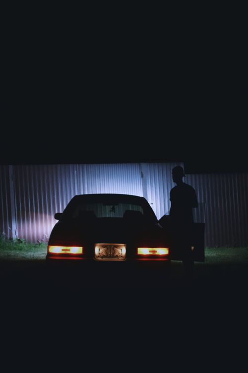Free Shadowy figure standing by a parked car under dim lighting, creating a mysterious urban scene. Stock Photo