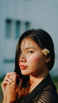 A beautiful young Asian woman poses outdoors, adorned with a flower in her hair, captured in natural light.