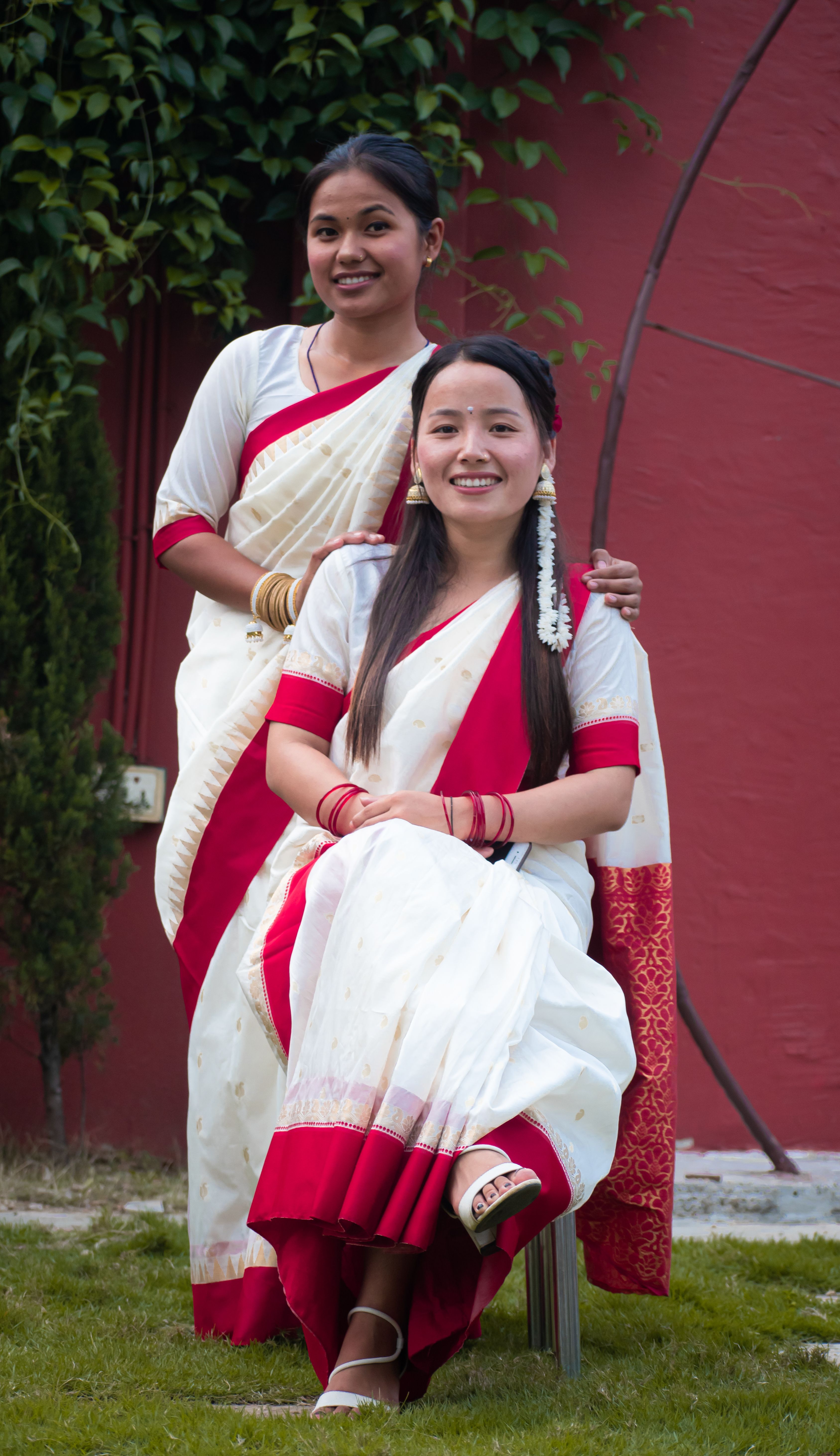 Two women in traditional Indian sarees, smiling outdoors against a natural background.