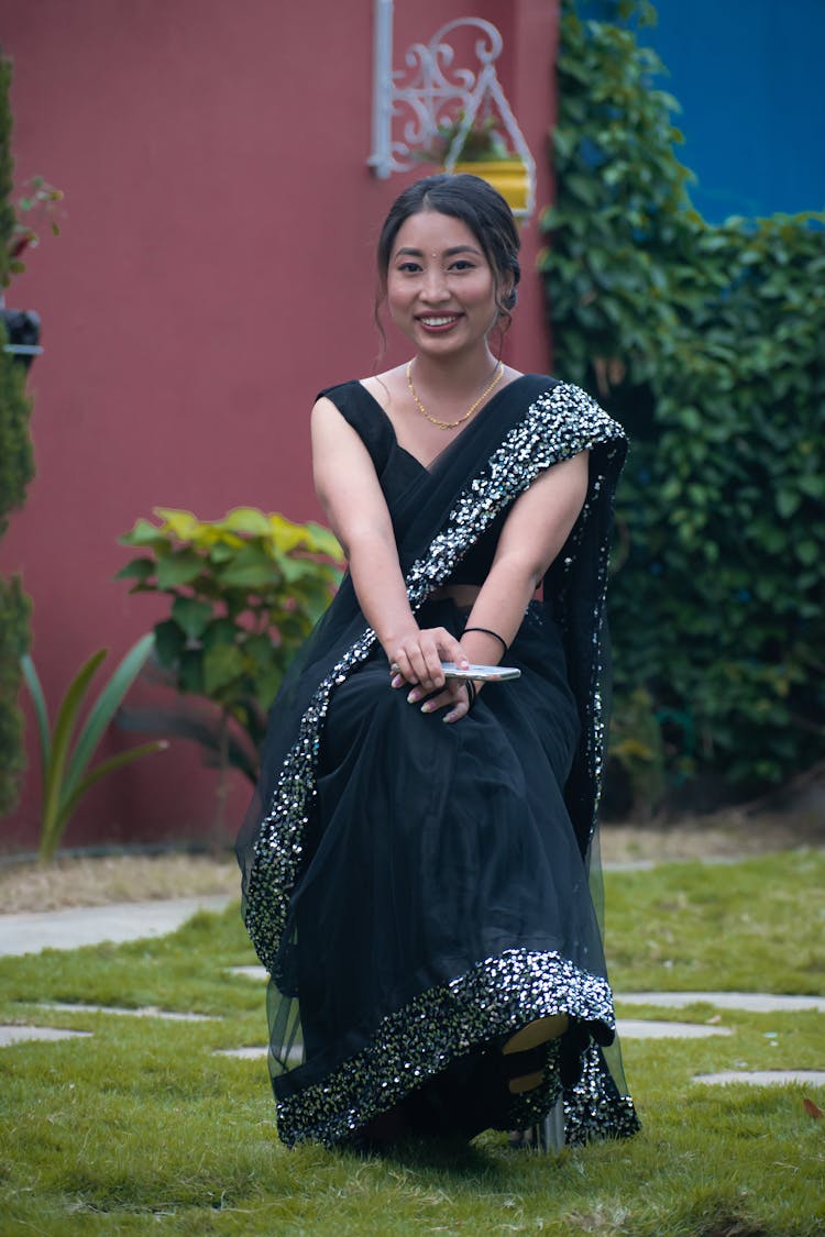 Photo Of A Smiling Woman Sitting In A Garden, Wearing A Black Sari And Keeping A Mobile In Hand