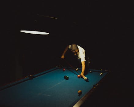 A focused player takes aim at a billiard table illuminated by overhead light.