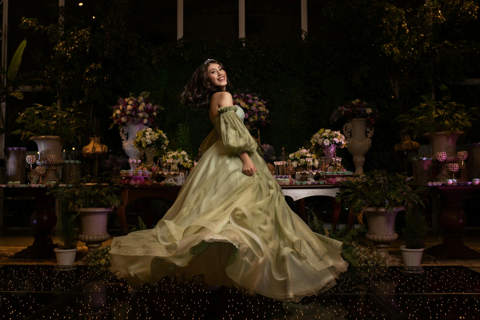 Elegant young woman in a flowing gown posing with a backdrop of lush florals and decorative table indoors.