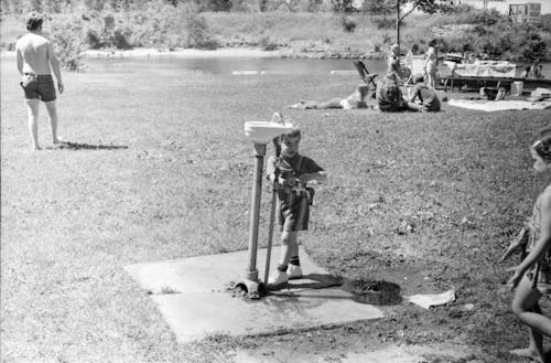 Grayscale Photo of a Kid Standing Beside a Drinking Fountain