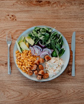 Delicious plate featuring avocado, corn, salad, sausages, and mashed potatoes on a wooden table.