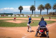 Children playing baseball in an outdoor field under a bright blue sky with palm trees in the background.