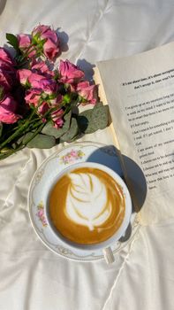 A latte with beautiful art, pink flowers, and an open book in soft natural light.