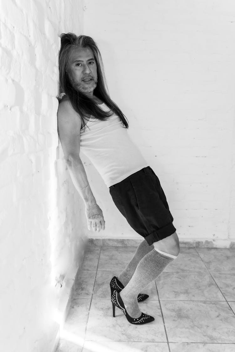 Man In White Tank Top Leaning Against A Concrete Wall Wearing High Heels