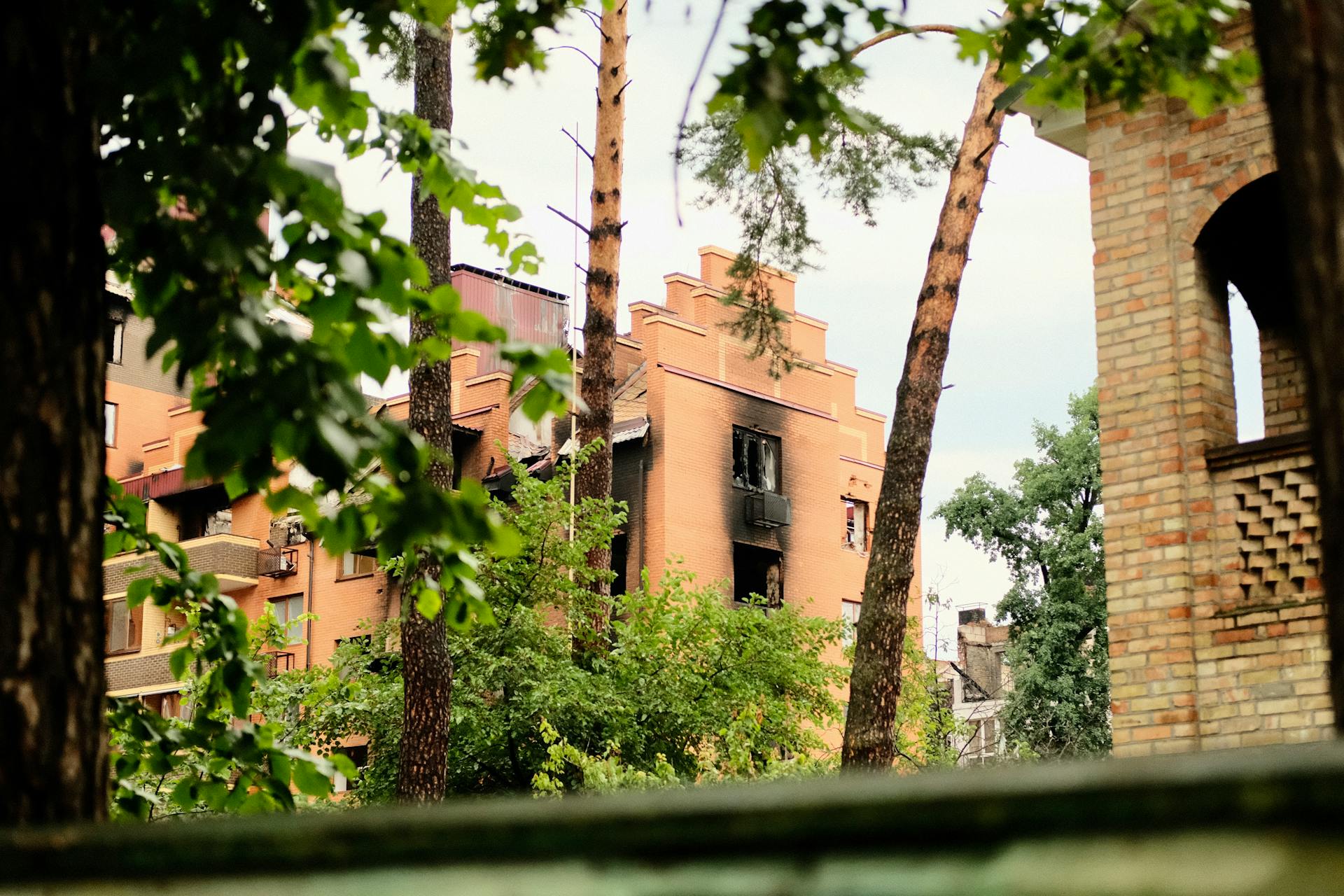 A damaged residential building, charred by fire, stands among lush trees in an urban setting.