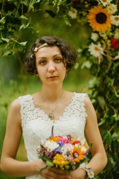 A bride in a lace dress holds a colorful bouquet under a floral arch.