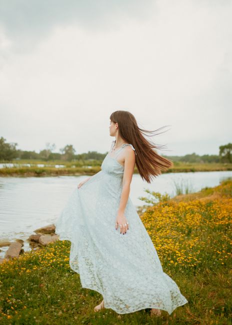 A woman in a lace dress gracefully spins by the riverbank surrounded by wildflowers.