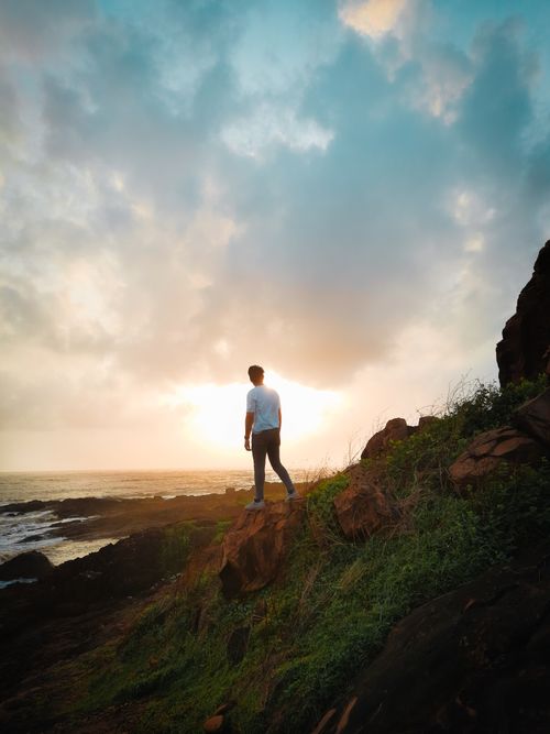 Free A lone person stands on a rocky coastal cliff, gazing at a stunning sunset over the ocean. Stock Photo