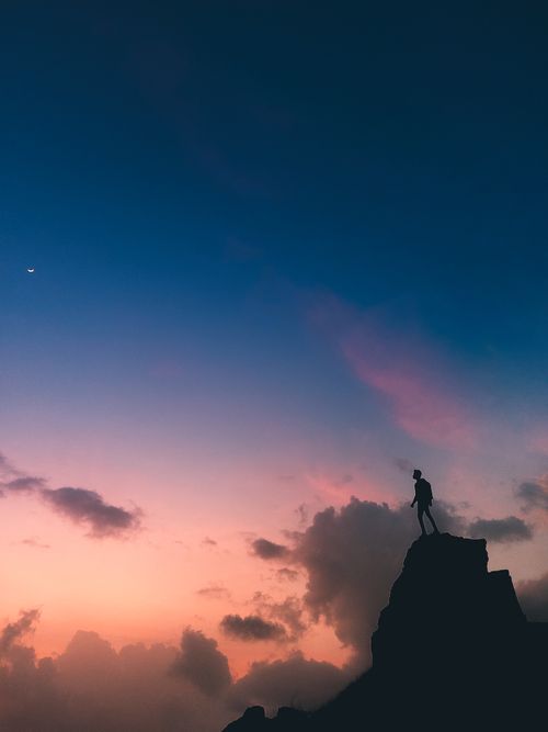 Free Silhouette of a man standing on a mountain peak with a colorful sunset sky, symbolizing achievement and tranquility. Stock Photo