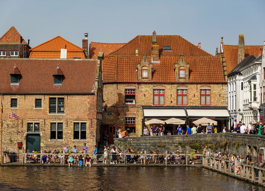 People enjoying sunny day by canal in historic Bruges with iconic brick architecture.