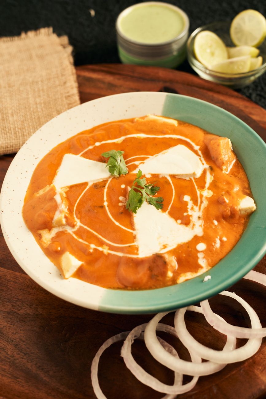 Rich, creamy orange-red paneer makhani curry in a dark bowl, dotted with tender white paneer cubes and garnished with bright green cilantro