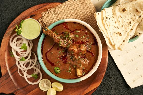 Bowl of red-brown lamb shank nihari with cilantro, onion rings, lime, green chutney and chapati
