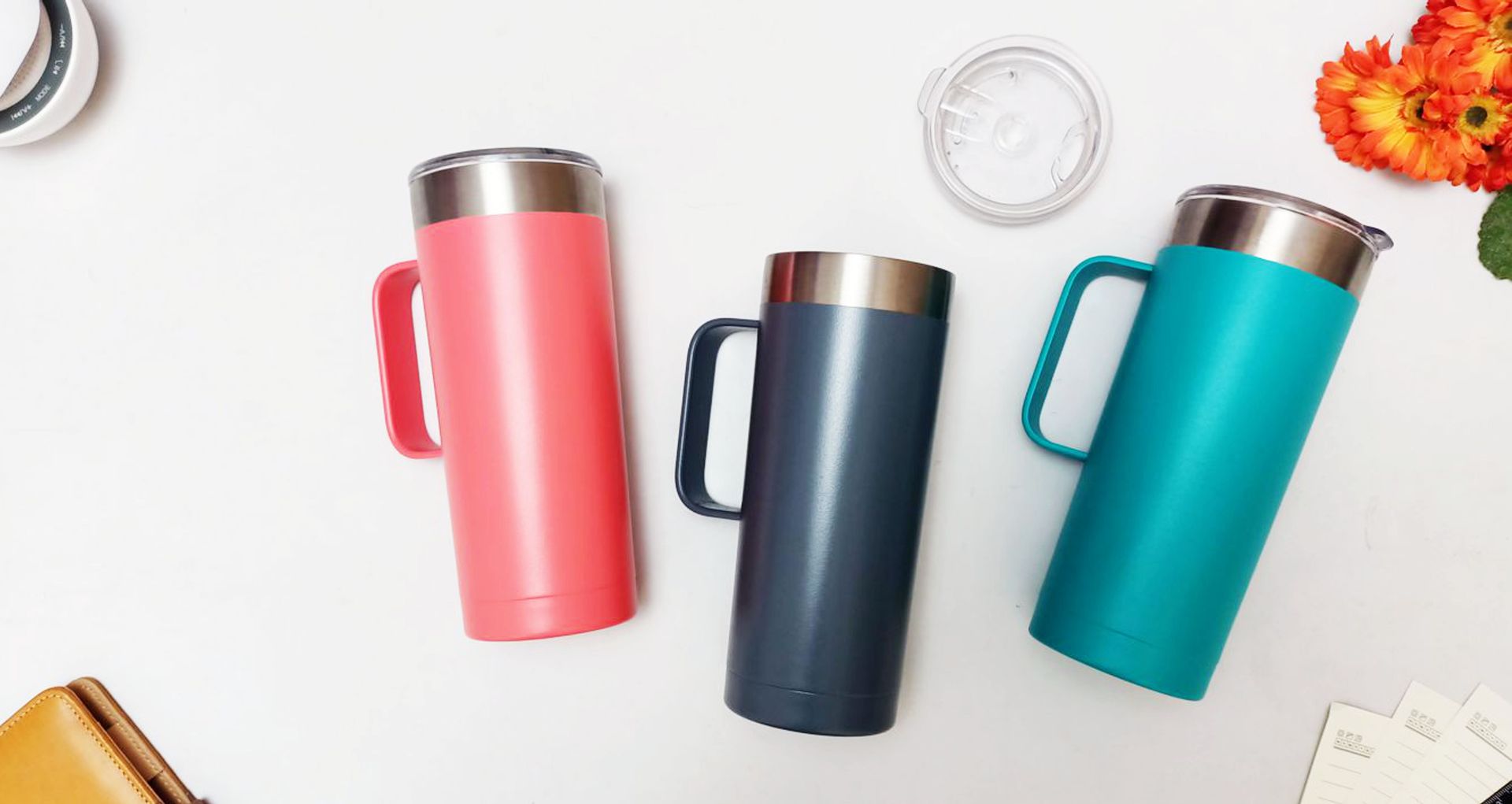 Three vibrant insulated travel mugs in pink, blue, and teal on a white surface with floral decoration.