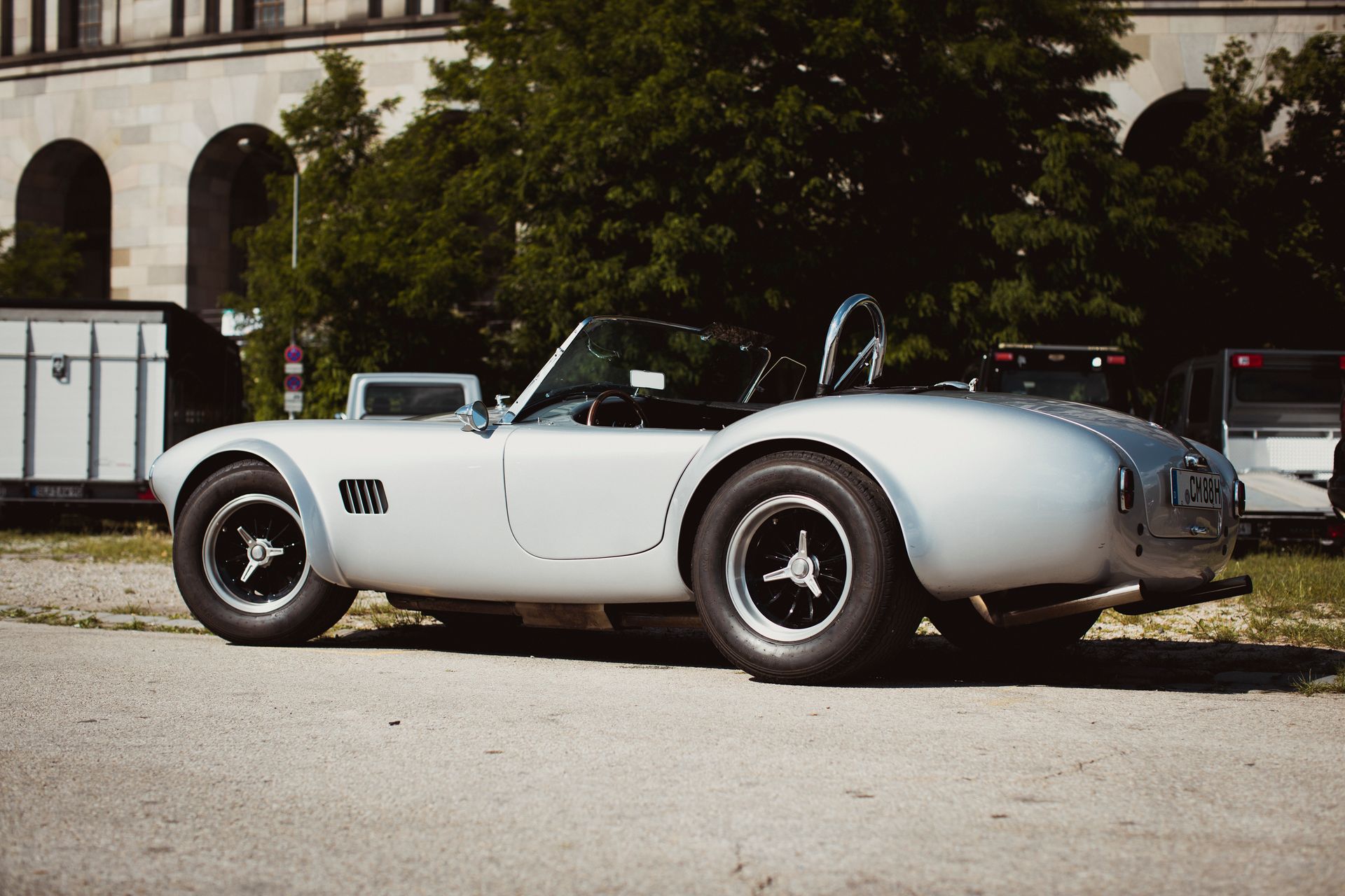 Classic AC Cobra sports car parked on a sunny day in an urban area with nearby trees and buildings.
