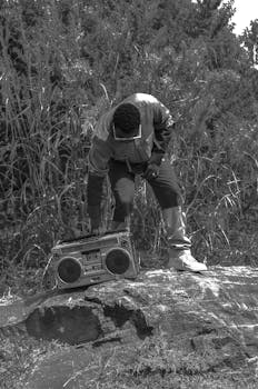 A man bends over a vintage cassette player outdoors in grayscale.