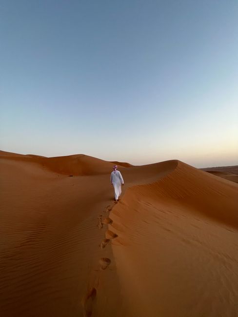 A solitary figure in traditional attire walking across desert sand dunes under a clear sky at sunset.