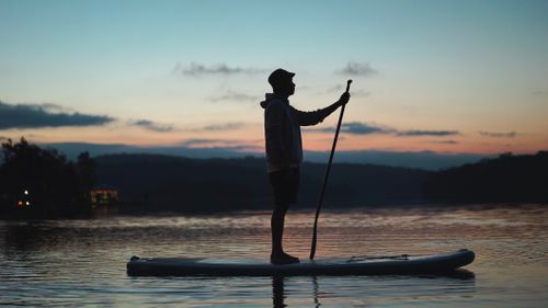 Free A person paddleboarding on a calm lake during sunset, creating a serene silhouette against the colorful sky. Stock Photo