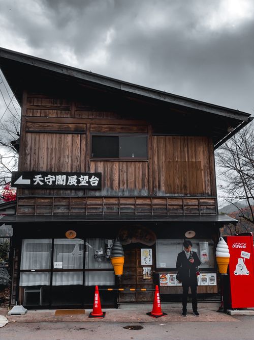 Free Man in black jacket stands by a vending machine near a traditional wooden house under a gloomy sky. Stock Photo