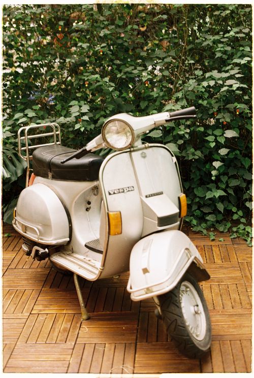 Free Classic white Vespa scooter parked on a wooden patio surrounded by lush greenery. Stock Photo