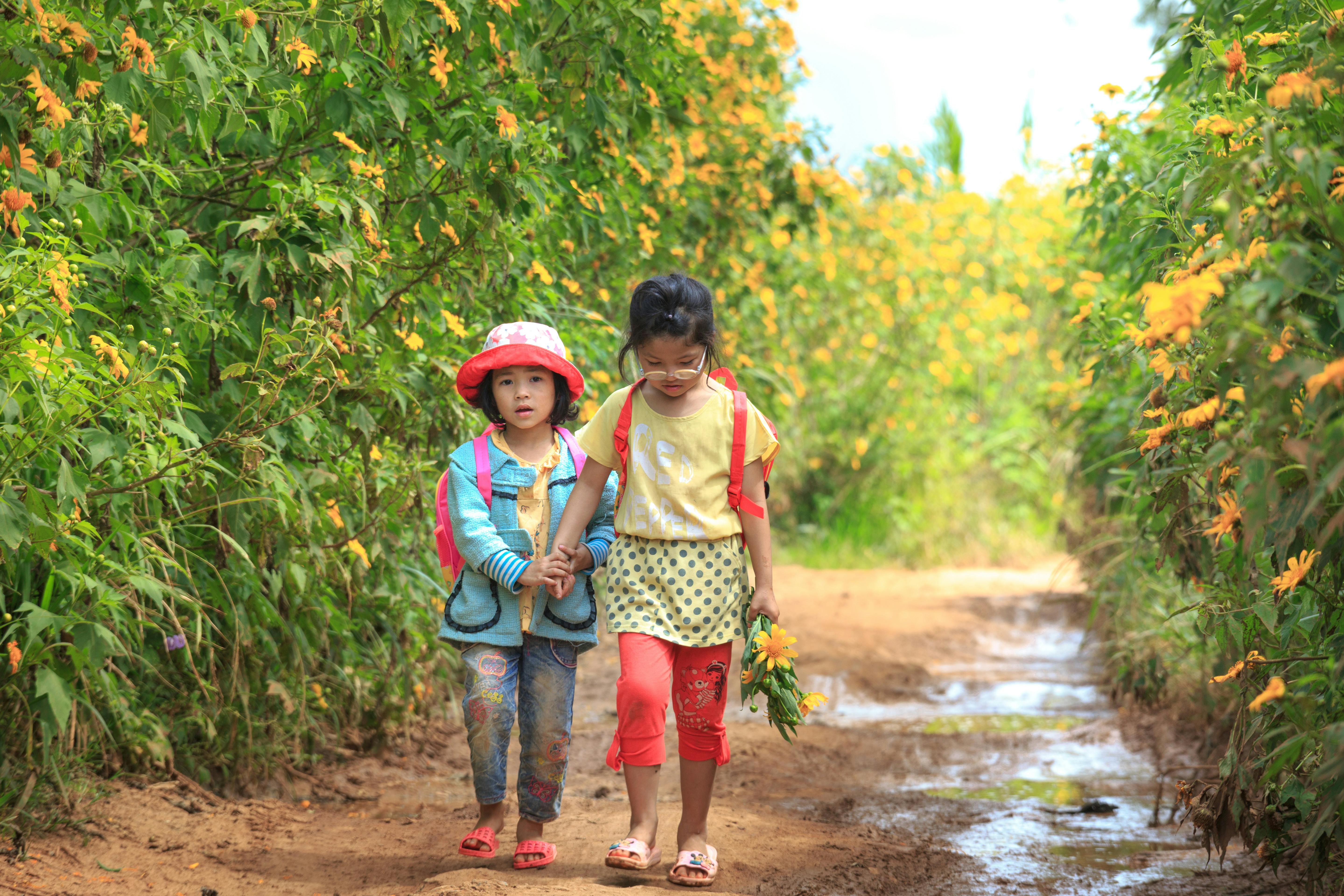 Two young girls walking together on a vibrant flower-lined path, holding hands joyfully.