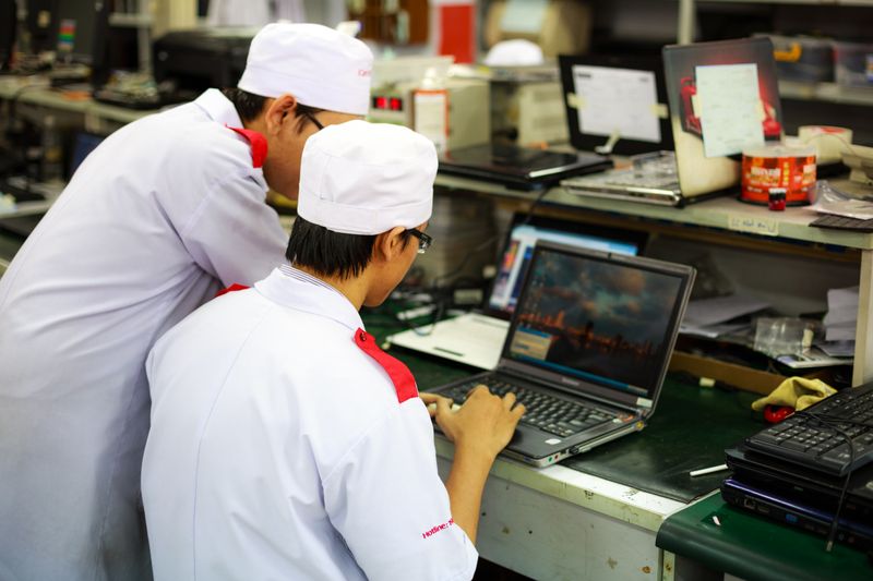 Two technicians in white coats and caps at a workshop bench, one working at an open laptop, with parts trays, tools and a second machine on the bench behind them