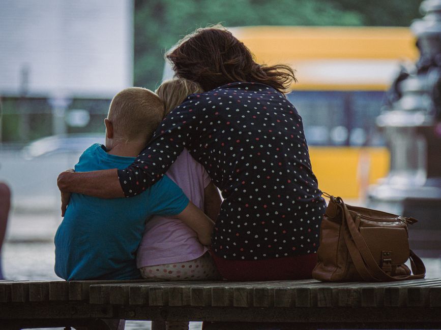 Una madre abrazando a sus dos hijos mientras están sentados en un banco de parque al aire libre.
