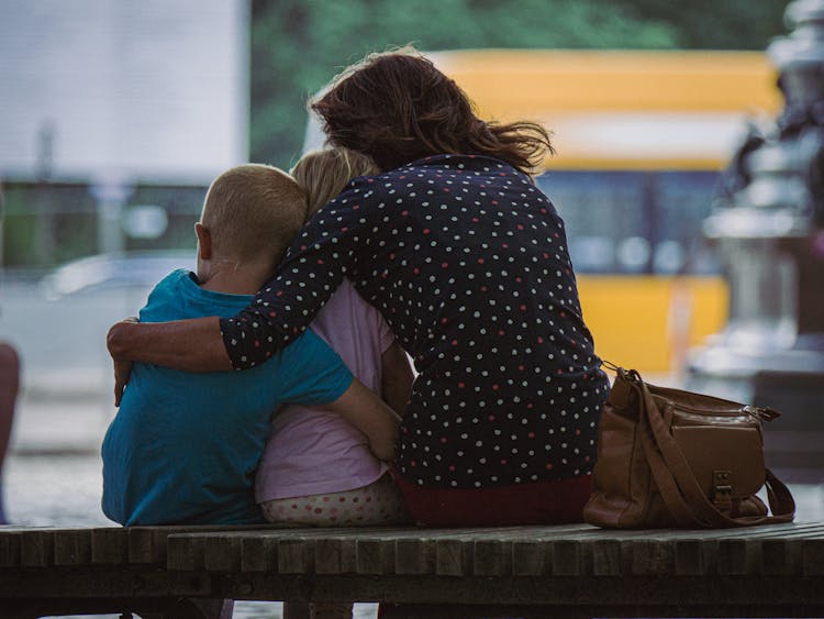 Back View Shot Of Mother And Kids Hugging Each Other While Sitting On A Wooden Bench