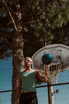 A young man holding a basketball in front of a hoop with a blue sky backdrop.