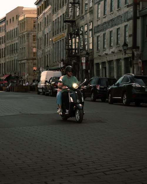 People Riding on a Black Scooter while Strolling on the Road Near Concrete Buildings